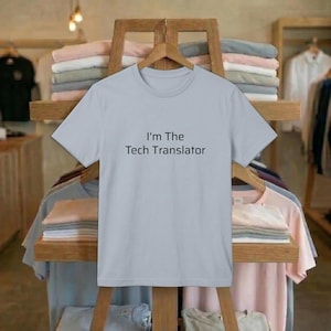 May include: A light blue t-shirt with the text "I'm The Tech Translator" in black. The shirt is hanging on a wooden rack with other folded shirts in the background. The t-shirt is a crew neck style.