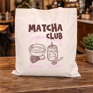 May include: A cream-coloured tote bag with the words "Matcha Sippin' Club" in brown and pink. The bag features illustrations of matcha preparation tools and a jar of matcha. The words "Good Mornings" and "Warm Matcha" are also printed on the bag.