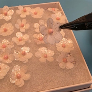 May include: A selection of delicate, white, glitter-dusted paper flowers with pink centres, arranged in a white box. The flowers are in a variety of sizes and shapes, suggesting a craft or decorative item. A black pen is touching one of the flowers.