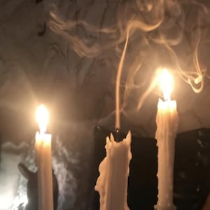 Könnte beinhalten: Drei brennende weiße Kerzen mit schmelzendem Wachs und aufsteigendem Rauch. Die Kerzen befinden sich in Glashaltern und auf einer dunklen Oberfläche. Die Flammen sind hell und die Szene ist schwach beleuchtet.