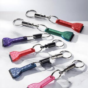 May include: A collection of keychains with a scraper design in various colors, including red, green, purple, blue, and pink. Each keychain has a silver metal ring and a black scraper blade. The keychains are arranged on a white surface.