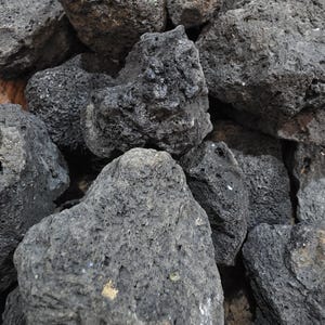 May include: A pile of dark gray lava rocks with rough, porous textures. The rocks vary in size and shape, with visible holes and crevices. The overall appearance is natural and rugged, with a slightly uneven surface.