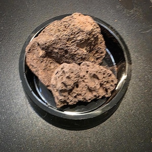 May include: Two pieces of porous, reddish-brown volcanic rock are resting inside a circular, glossy black plastic tray. The rocks feature a rough, uneven surface with numerous small holes and crevices throughout their entire structure.