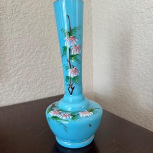 May include: A light blue glass bud vase with a slender neck and a rounded base. The vase features hand-painted floral motifs in shades of pink and white with green leaves and brown stems, extending from the base up the neck of the vessel.