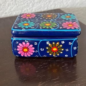 May include: A rectangular dark blue box with a lid featuring hand-painted floral patterns in pink, turquoise, green, and brown. The sides of the box are decorated with similar flower motifs and small white dots, outlined by thin light blue lines.