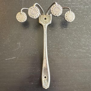 May include: A silver-toned metal wall hook featuring a vertical handle base with two mounting holes. The top section branches into four curved arms, each ending in a circular decorative knob embossed with a geometric sunburst pattern.