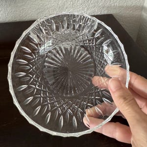 May include: A clear glass bowl featuring a detailed pressed pattern with a central starburst design surrounded by radiating leaf-like motifs and a cross-hatched diamond texture along the upper rim, creating a reflective and faceted appearance.
