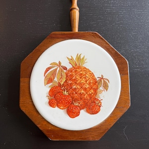 Puede incluir: Un salvamanteles octogonal de madera con un azulejo de cerámica blanco circular en el centro. El azulejo muestra una colorida ilustración de una piña grande rodeada de varias fresas rojas y hojas verdes. Un mango de madera sobresale del borde superior.