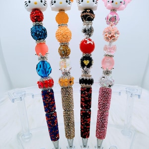 Pode incluir: Quatro canetas decorativas com toppers de personagens, incluindo Hello Kitty e My Melody. Cada caneta apresenta um esquema de cores diferente de contas brilhantes e strass. Os barris das canetas são cobertos com strass correspondentes.