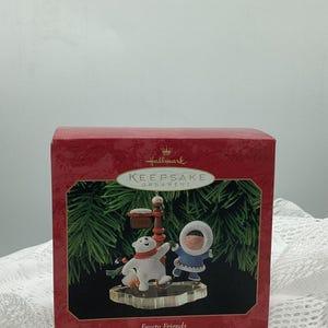 Può includere: Una scatola di ornamenti Hallmark Keepsake della serie da collezione Frosty Friends del 1999. La scatola mostra un orso polare bianco e una figura in parka blu che pattinano vicino a un cartello con la scritta 1999 su uno sfondo di pino verde.