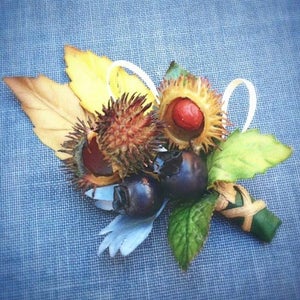 May include: A floral arrangement featuring a cluster of brown and yellow spiky seed pods, two dark purple berries, and green leaves. The arrangement is tied together with a brown ribbon.