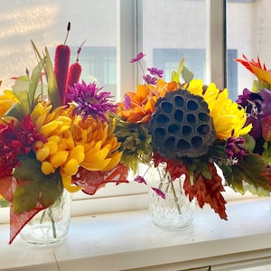 May include: Five glass jars filled with fall-themed floral arrangements. The arrangements feature yellow, red, and purple flowers, along with brown seed pods and green leaves.