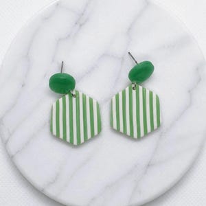 May include: A pair of dangle earrings featuring a small, smooth, solid green oval stud connected to a larger, flat, hexagonal pendant. The hexagonal pendants are decorated with a repeating pattern of vertical white and green stripes.