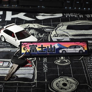 May include: A white toy car and a keychain with Japanese text and a mountain scene. The keychain has a car graphic. The items are on a black surface with car diagrams and text.