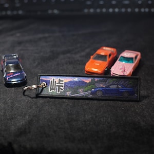 May include: A collection of miniature toy cars and a keychain. The keychain features a scenic image of a mountain and a blue car on a road, with Japanese text. The toy cars are in shades of blue, orange, and pink.