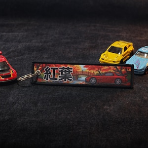 May include: A red toy car and three other toy cars are displayed on a dark gray surface. A keychain with Japanese text and a car graphic is also present. The keychain features a bridge and autumn foliage.