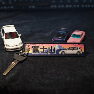 Puede incluir: Un coche de juguete blanco con interior rojo está junto a un llavero con texto japonés y un gráfico de coche. Otros dos coches de juguete, uno azul y otro rosa, están en el fondo. El llavero tiene un gráfico de montaña.