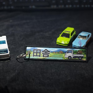 Peut inclure: Un ensemble de petites voitures miniatures et un porte-clés. Le porte-clés présente du texte japonais et une scène de paysage avec un petit camion. Deux voitures sont vert citron et bleu clair. Un camion jouet blanc et argenté est également présent.