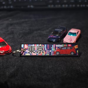 May include: A red toy car and three other miniature cars are arranged on a dark surface. A rectangular keychain with the word "Tokyo" in Japanese and a red car graphic is in the foreground. The keychain has a colorful cityscape design.