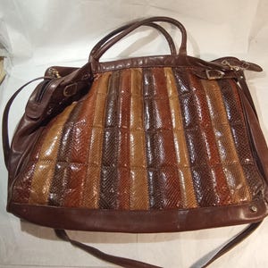 Può includere: Una borsa grande caratterizzata da un pannello testurizzato con motivo a pelle di serpente multicolore nelle tonalità marrone, beige e cioccolato fondente. La borsa include finiture in pelle marrone scuro, due manici, una tracolla lunga e dettagli in metallo.