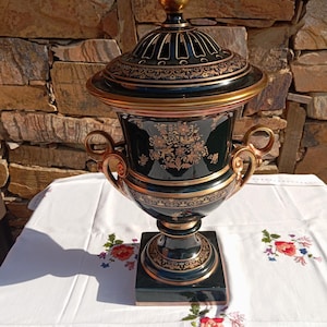 May include: A black ceramic urn with a matching lid, featuring intricate gold floral patterns and scrollwork. The vessel has two ornate side handles, a pedestal base, and a gold-toned finial on top, showcasing a glossy finish throughout.