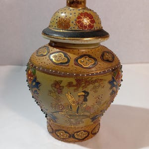 Puede incluir: Un jarrón de cerámica decorativo con tapa y remate dorado. El jarrón presenta un fondo beige con diseños florales y de nubes dorados, rojos y azules. En el cuerpo del jarrón se aprecia una escena pintada de una persona.