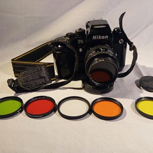 Peut inclure: Un appareil photo Nikon F4 noir avec un objectif et une sangle, ainsi que plusieurs filtres d'objectif colorés. Les filtres sont verts, rouges, blancs, oranges et jaunes. L'appareil photo est sur une surface blanche.