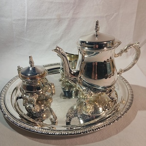 Op de afbeelding: Een zilveren theeservies op een rond dienblad. De set bevat een theepot, een melkkan, een suikerpot met deksel en een dienblad met een decoratieve rand. De theepot heeft een gebogen handvat en een decoratieve knop op het deksel.