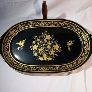 Op de afbeelding: Een ovale zwarte serveerschaal met gouden bloemen- en krulwerkontwerpen. De schaal heeft een gouden rand en handvatten, en een bruin handvat aan de bovenkant. Het midden van de schaal heeft een gouden bloemstuk.