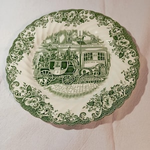 May include: A white ceramic plate with a green floral border and a central illustration of a horse-drawn carriage in front of a building. The plate has a scalloped edge and the words "COACHES DAILY" are visible.