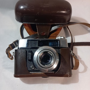 Może przedstawiać: Aparat Voigtlander Color-Lantar 2.8/50 z obiektywem Prontor 300 LK, posiadający srebrno-czarny metalowy korpus umieszczony w brązowym skórzanym etui ochronnym z dopasowanym skórzanym paskiem na szyję i otwieraną górną pokrywą.