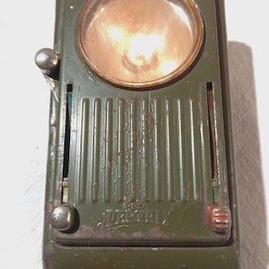 May include: Vintage olive green flashlight with a circular lens and a metal casing. The flashlight has a switch on the side and a red accent on the bottom. The word "DELTRON" is printed on the front.