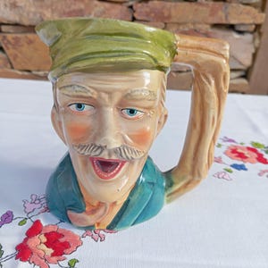 Peut inclure: Une cruche en céramique en forme de personnage portant une casquette verte et une veste bleu sarcelle foncé. La figurine a une moustache, des yeux bleus et une bouche ouverte. L'anse est texturée comme du bois et la finition est brillante.