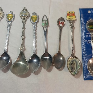 May include: A collection of seven silver-plated souvenir spoons featuring decorative handles with various colourful emblems and crests. One spoon remains sealed in its original blue plastic packaging labelled Souvenir of Canada Silverplated.