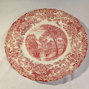 May include: A cream-coloured plate featuring a red floral border and a central landscape scene. The landscape shows a building near a waterfall and trees. The plate has a scalloped edge.