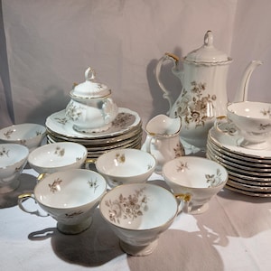 Puede incluir: Un juego de té de porcelana blanca con detalles dorados y motivos florales. El juego incluye una tetera, azucarero, jarrita para la leche, tazas, platillos y cuencos pequeños. Las tazas tienen asas doradas.