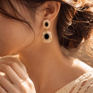 May include: Cream-colored drop earrings with a black oval center stone and gold-colored trim. The earrings have a circular top and a larger, rectangular bottom. The design features intricate detailing. The earrings are worn on the ear.