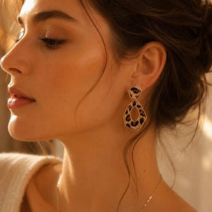 May include: Gold-coloured earrings featuring a leopard print pattern. The earrings have a geometric design, with a smaller top and a larger, open-centre bottom. A fine gold necklace is also visible.