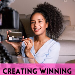 May include: A woman holding a makeup palette and brush, smiling in front of a video camera. The image has the text "CREATING WINNING VIDEOS" in a pink banner at the bottom. The woman is wearing a light blue top.