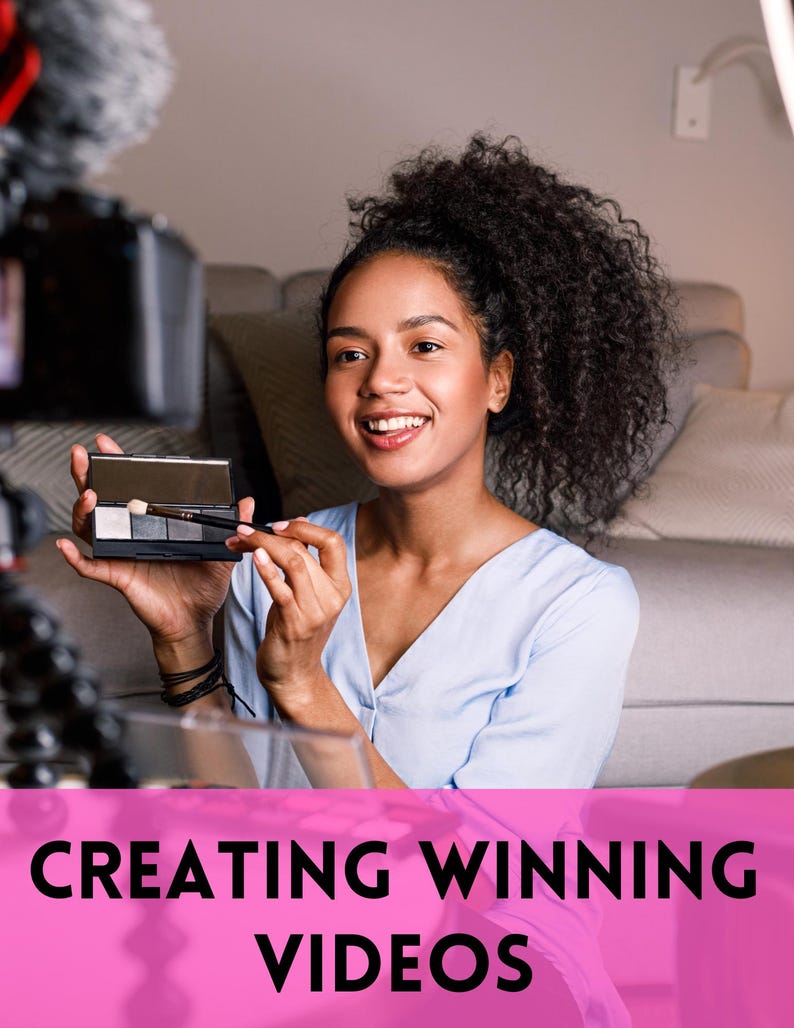 May include: A woman holding a makeup palette and brush, smiling in front of a video camera. The image has the text "CREATING WINNING VIDEOS" in a pink banner at the bottom. The woman is wearing a light blue top.