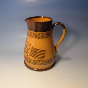May include: A tan-colored pitcher with a dark brown rim, handle, and base. The pitcher features an American flag and text, with decorative floral patterns. The design is reminiscent of vintage or antique pottery.