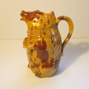 May include: A brown and yellow ceramic pitcher shaped like a pig. The pitcher has a brown glaze with a mottled pattern.