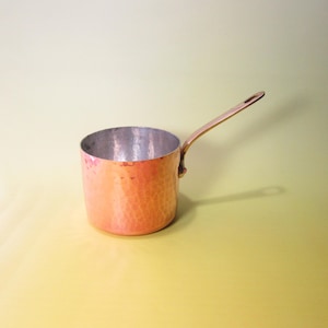 May include: A copper saucepan with a long handle. The pot has a hammered texture on the exterior and a shiny interior. The handle is attached to the side of the pot. The saucepan is sitting on a yellow surface.