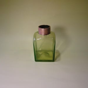 May include: A square, green glass bottle with a copper-colored rim. The bottle has a clear, transparent appearance and a simple, classic design. The bottle is empty.