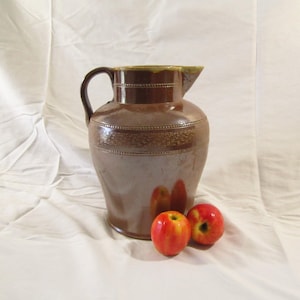 May include: A brown ceramic pitcher with a handle and a spout, decorated with horizontal bands and a textured pattern. Two red and yellow apples sit in front of the pitcher. The pitcher is approximately 12 inches tall.