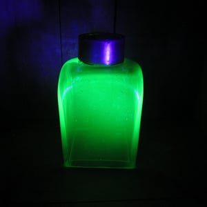 May include: A glowing green glass bottle with a dark purple cap. The bottle is rectangular with rounded corners, and the green liquid inside glows brightly against the dark background. The cap is a dark purple color.