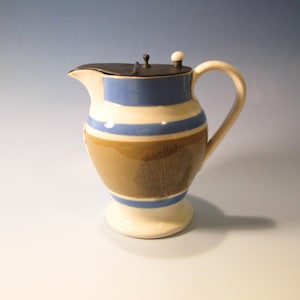 May include: A ceramic pitcher with a dark brown body, cream-colored base, and a cream-colored handle. The pitcher features blue and cream-colored horizontal stripes and a dark lid with a small knob. The spout is white.