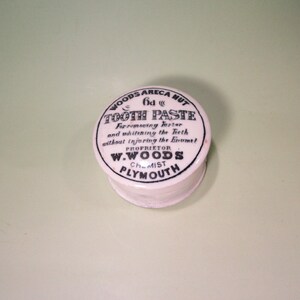 May include: A vintage white cylindrical container of "Tooth Paste" with black text. The label reads "Woods Areca Nut" and "For removing tartar and whitening the teeth without injuring the Enamel." The proprietor is W. Woods, Chemist, Plymouth.