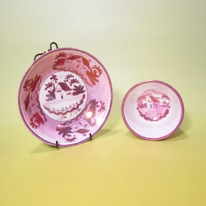 May include: Two pink and white ceramic bowls. The larger bowl is displayed on a black metal stand. Both bowls feature a landscape design with a house, trees, and a fence. The bowls have a pink rim and a pink and white interior.