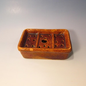 May include: A rectangular, brown ceramic soap dish with a glossy finish. The dish has three sections, each with several small, round drainage holes. A larger central hole is also present. The soap dish is designed to hold and drain soap.